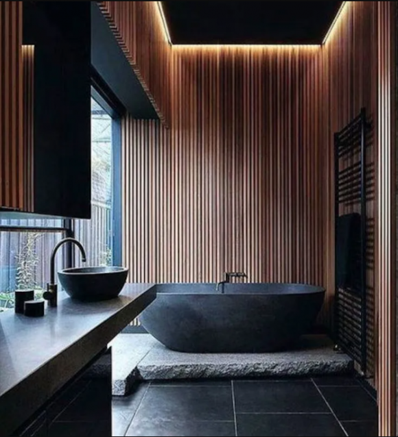 A stone freestanding bath in the center of an open space, on a grey stone slab, on top of black oversized tiles with grey grout. A matching basin bowl sits on the counter with faucet. Wooden slats cover all the vertical surfaces, giving the space a warm feeling.