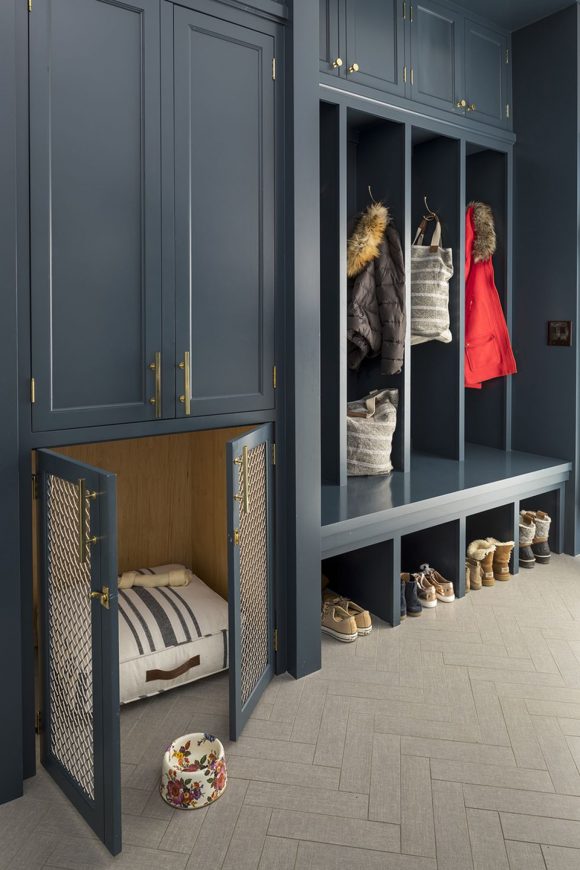 Beige-grey herringbone wood floors underneath dark cabinetry featuring a brass metal grill-fronted 2-door dog crate, next to 4 cubbies for shoes, 4 tall spaces for coats, hats, and scarves with hooks, and brass handles.