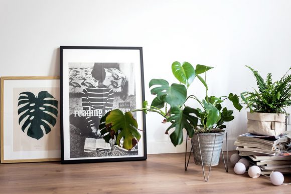 A tan-framed image of a plant frond sits on the floor next to a black-and-white-image of a woman reading, next to a metal-potted plant also on the floor, and a stack of magazines topped with a fern next to that.