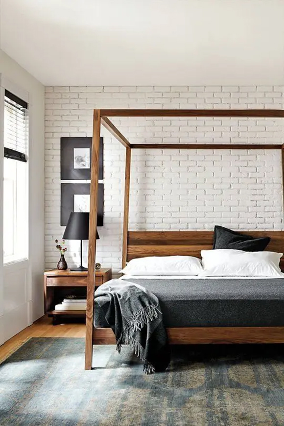 A simple wooden canopy frame in front of a painted brick wall, with black framed images, a black bedside table lamp, and grey blanket with white sheets.