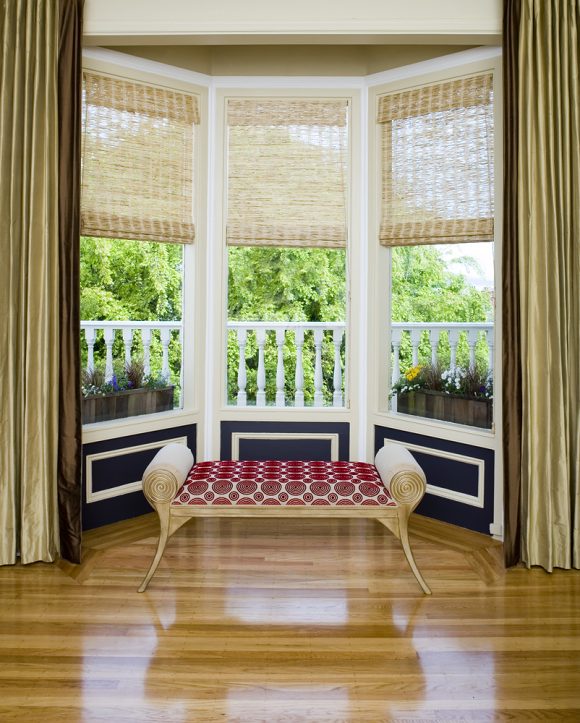 Bay window showing green plants outside with stacked gold draperies from floor-to-ceiling at the sides of the window. Woven grass blinds are extended half-way down the windows. A bench seat of rolled gold arms upholstered with a red circular pattern sits in front of 3 dark blue and white rectangular wood architectural details under the windows. The floor is a reddish shiny hardwood.