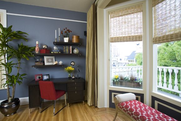 Corner of a living with blue walls and a white picture rail near the ceiling. A desk with open shelving above sits near a bay window. Gold floor-to-ceiling draperies are stacked at the side of the window, while woven grass blinds are extended half-way down the windows. A gold bench seat with scrolled arms is upholstered in a red circular pattern to create another seating area.