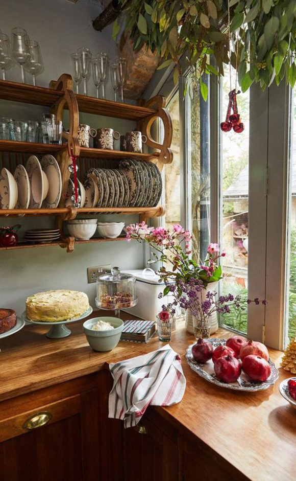 A homey kitchen with wood countertops filled with fruits and cakestands. Open shelving has slats for plates and bowls, with glassware above. A sprig of purple flowers from the garden complement the green view outside the window. A striped towel is scrunched in the foreground, as if someone just walked away.