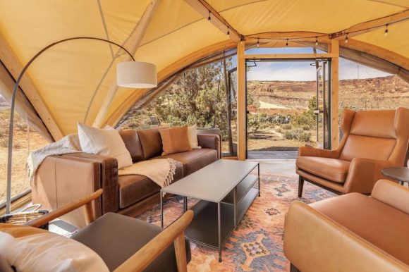 A yellowish canvas tent arches over an entire living room suite of leather sofa and 3 chairs, with a metal coffee table in the center sitting atop a Southwest themed orange and blue rug. The arched entrance folded up reveals a beautiful view into the green and dusty desert.