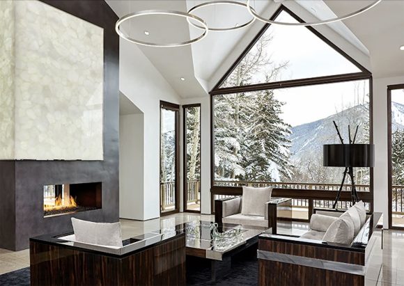 Aspen living room with high ceilings, a triangular shaped window with a view out to the snowy mountains, a triple ring pendant light hanging over a living room seating area with dark and reddish brown sofa and armchairs with white cushions, in front of a large ribbon fireplace under a dark brown wall with a white patterned square above. The walls and ceilings are painted white.