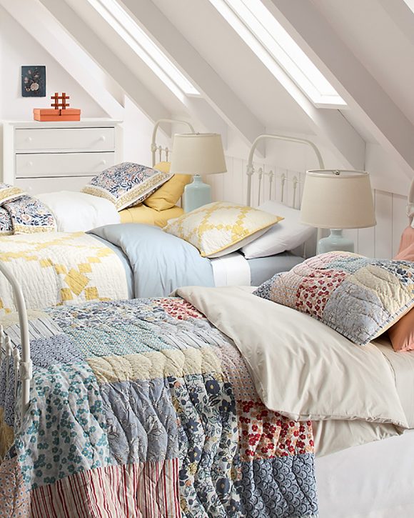 Under a white slanted ceiling with three skylights, 3 beds with rounded metal frame headboards are covered in quilts with multiple patterns and colors of fabric. Two matching lamps sit between the beds on beside tables, and a white chest of drawers is visible in the background.