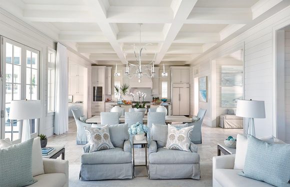 Light colored room painted in white, with robin's eggshell blue furniture and pillows arranged in conversation groups, a dining table in the center and cream cabinetry in the kitchen at the rear of the space.