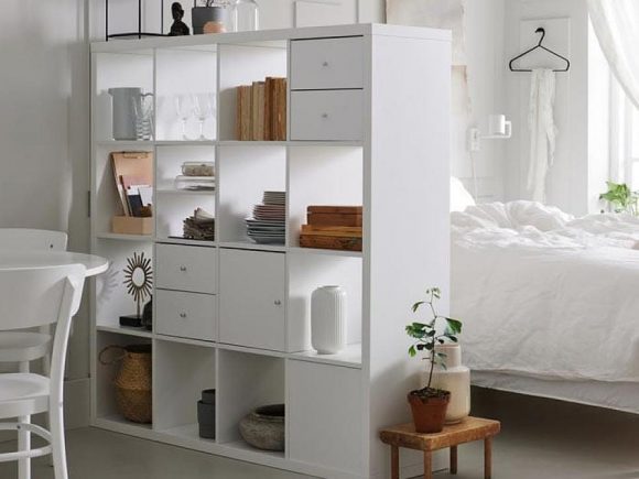 White open bookshelf has drawers and doors with silver pulls, while books, plates, and glassware adorn the open storage spaces. On the left, a white table and chairs. On the right, a bed with white linens.