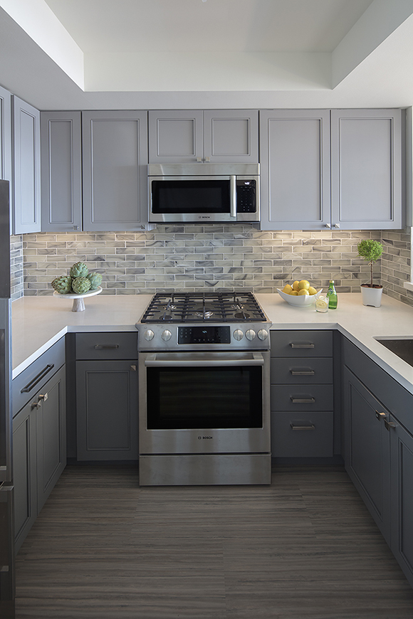 A compact kitchen in 2 shades of grey cabinetry with the darker shade on the bottom cabinets, plus off-white countertops, and a subway-tiled backsplash with multiple shades of grey and off-white tiles. The stove and microwave above it are silver and black, and the counters are dressed with a plate of green vegetables, a bowl of lemons, a green bottle of spring water, and a small green plant.