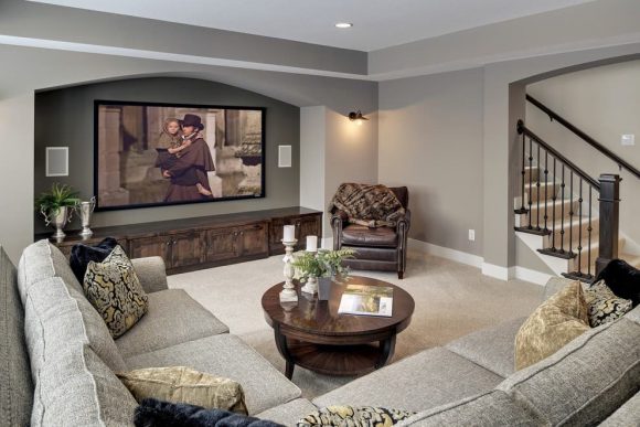 A greyish-tan traditional-style living room with textured corner sofa in tan and white colors plus multiple patterned throw pillows in gold, black, and white. A round dark wood coffee table sits in front of the sofa, with a dark brown leather recliner opposite. Near the leather recliner is a large projection screen with built-in speakers either side, and a low wall of cabinetry in the front, forming a black counter surface on top. A sconce downlight is visible on one wall, with recessed lighting coming from the ceiling. To the right, a set of stairs going up is partially in view, with metal railings. The tables and counter surfaces are dressed with white candles on tall tan candleholders, and plants in white Grecian vases.