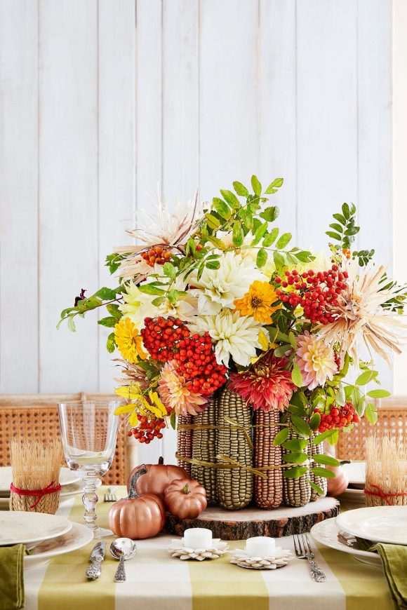 White painted wooden slats on the walls behind are the backdrop to a dining table covered with a gold and white large checked tablecloth set with white plates, clear glasses, and silverware. 

A bold centerpiece is created by tying together gold and copper painted ears of corn standing on their ends, filled with fresh flowers in red, white, orange, pink, yellow, and plenty of greenery.