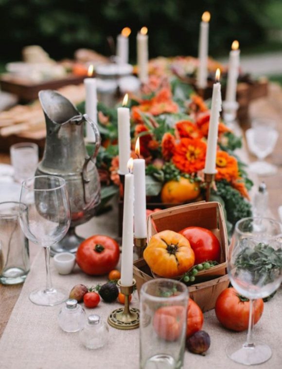 A clustered table of clear glasses, silver pitcher, white candles in brass candle holders, and wooden square boxes overflowing with orange and red peppers, tomatoes, apples, and other fruits and vegetables, along with orange flowers, and green plants out of focus in the background.
