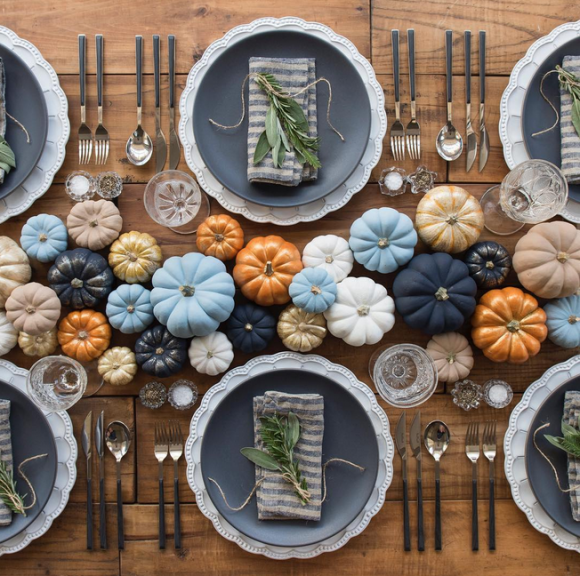 Overhead view of a Thanksgiving dining table with grey plates on top of bigger white plates with scalloped edges, and grey and white striped cloth napkins folded under springs of greenery. Silverware is set either side with 2 forks, 2 knives, and a spoon for each place setting, plus a water glass. 

In the center of the table are 29 small pumpkins in multiple sizes, painted varying shades of solid navy blue, solid sky blue, solid gold, solid white, solid tan, one in tan, gold, and orange stripes, and a solid shiny orange color. All are set upon a plain exposed wood plank table.