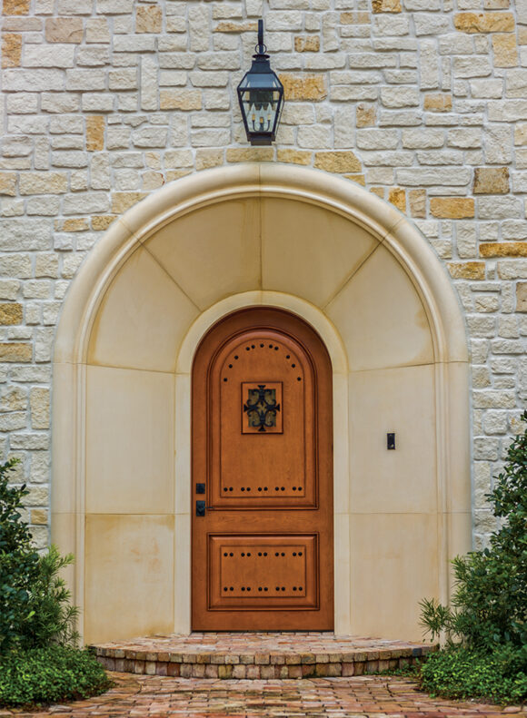A large off-white stone arch surrounds a reddish wood arched front door, studded with metalwork. The brickwork around the stone is off-white and yellowish in tone. A single brick step leads to the door.