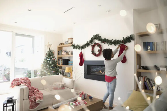 A woman of color hanging a Christmas stocking from a pine garland above a mantle-less ribbon fireplace in a light-colored living room with light streaming in the side window.