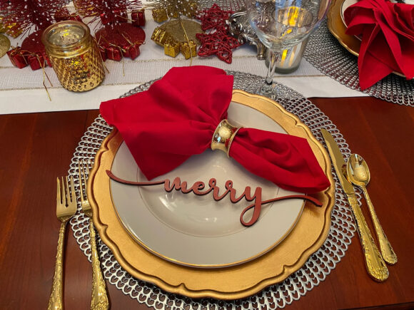 Holiday table with a large red fabric napkin in a gold ring laid above a wooden laser printed word 'merry' atop a white plate, on top of a gold charger, layered on top of a silver place setting, with gold tableware either side, and glasses and candles above.