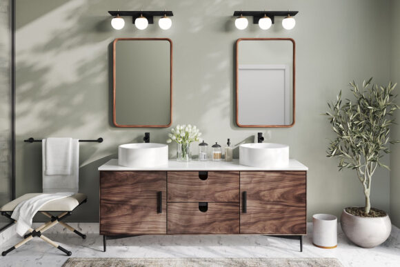A bathroom vanity in natural wood finish fronted cabinetry, with a pair of round white vessel slinks below matching mirrors on the wall, below matching triple sconce lights. A plant sits at the right of the space with a folding seat and white towel at the left.
