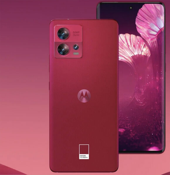 A Motorola cell phone in Viva Magenta with 2 cameras and a flash features the Pantone color chip logo at the bottom, below the Motorola logo in the center. Behind, a phone case bears an illustration of shapes in colors like pink, fuscia, and of course, Viva Magenta.