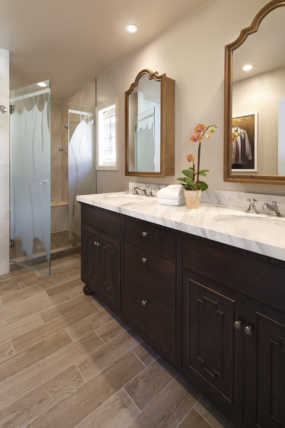 A partly open frosted shower door at the far left of the image, while in the foreground, a double-sink marble countertop over dark wood cabinetry, with a pair of mirrored medicine cabinets, above varied wood-look floor tiling. Recessed ceiling lights shine down on the bathroom as a whole.