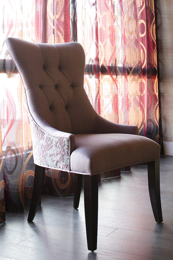 Close up of a purple tufted head chair with raised velvet floral patterning on the outside of the chair, while the seat and back have a light purple velveteen fabric. The chair sits in front of a window with gauzy curtains in orange and hot pink geometric patterns. The flooring is a dark wood.