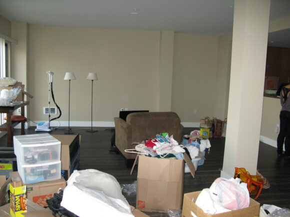 BEFORE: A blank, beige wall, boxes of clothes and personal belongings, floor lamps, table, and a small sofa fill the dining area of an Oakland condo.