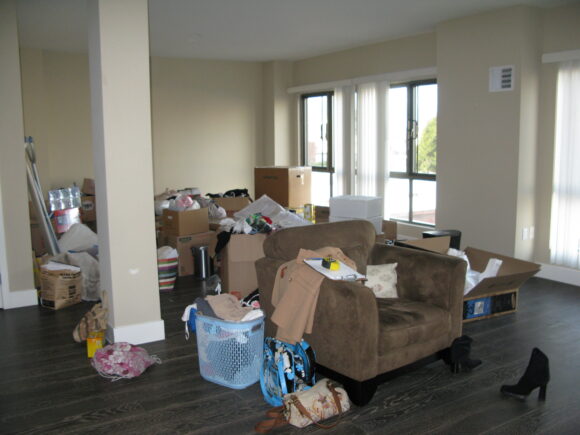 A brown oversized armchair sits in front of a pile of boxes, containers, laundry bins, bags and personal items. A large support column is seen at the left side of the space. To the right are a pair of windows with flat black frames and gauzy white curtains.