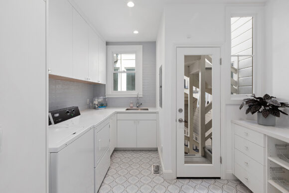 A mostly-white laundry and mudroom, with a backsplash of pale blue subway tiles and circular patterns on porcelain floor tiles. The white washer and dryer under a white countertop with upper and lower cabinets are closest to the viewer, with a sink and window on the far end wall. A mirror adorns the back of a small closet door, and a plant sits on the counter. Two windows allow light into the space. Everything except the plant is white or pale blue.