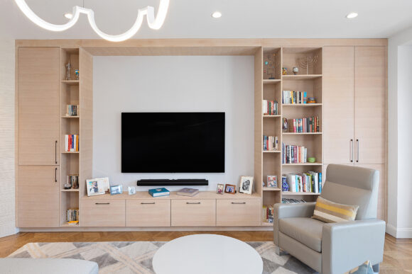 Under the modern chandelier, and against the rear is a wall of custom cabinetry in pale peach shades. Closed cabinets flank open shelving with deep drawers at the bottom. A television hangs on the wall in the center. The greige chair is to the right, with the white-topped coffee table in front of it, on top of the diamond-shape patterned rug in grey and peach.