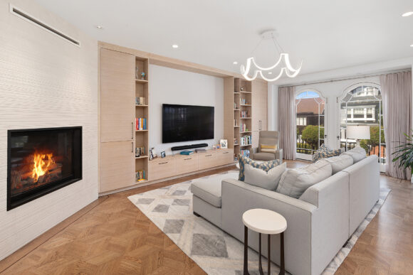 Facing the other direction in the same living room space, we see the sofa from the back, facing the TV, media center, and shelving full of books. A fireplace is visible at the left wall angled towards the viewer. A light brown wood flooring has geometric patterns, and a rug with diamond patterns anchors the seating area.