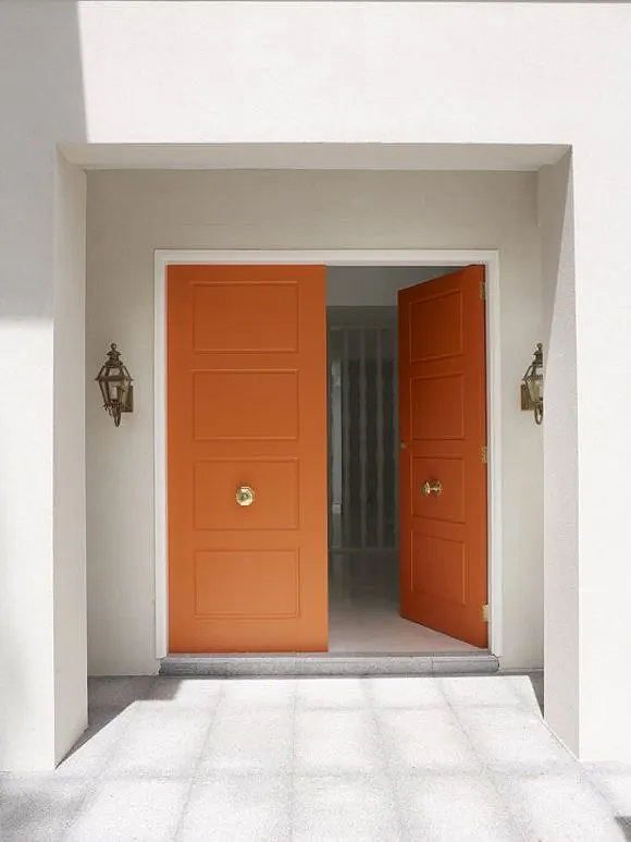 Bright orange front double doors with gold center door pulls. The right door is open, inviting, and two classic gold wall sconces that look like candle-carriers are hung either side of the doorway.