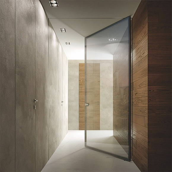 A long hallway with cement walls on the left side, wood walls on the right side, and a transparent glass pivot door outlined in black metal between you and the hallway end wall, which features a cement wall with a wood insert panel, combining the two materials simply and beautifully.