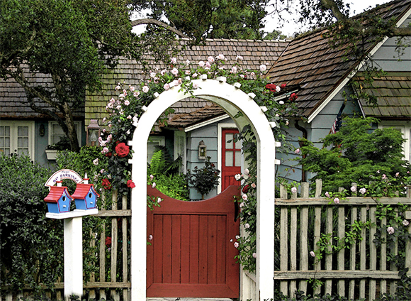 A light blue cottage with an arched white front gate entrance and a red gate, with pink flowers growing over the arch. A pair of blue birdhouses with red roofs form the mailboxes, labeled with the family's name are at the front.