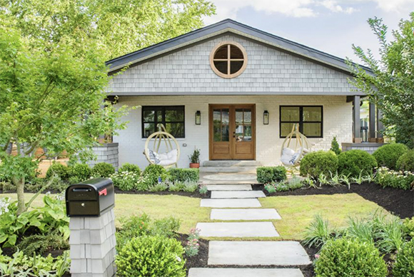 Craftsman home with light grey shingles across a pointed roof with a circle center window above the front door. The shingles are repeated on the mailbox support. Alternating pavers lead to the front porch and door with green plantings all around.