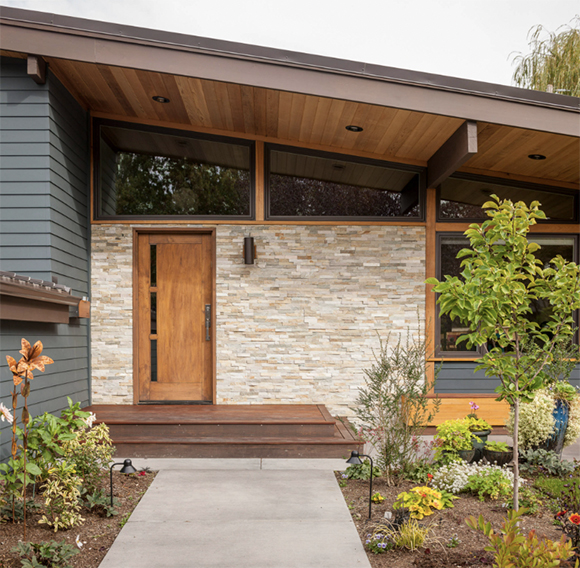 A midcentury modern exterior appeals with stonework on the front wall around the door, exposed wood beams around the windows and below the ceiling, and neatly trimmed natural plantings surround the approach to the front door.