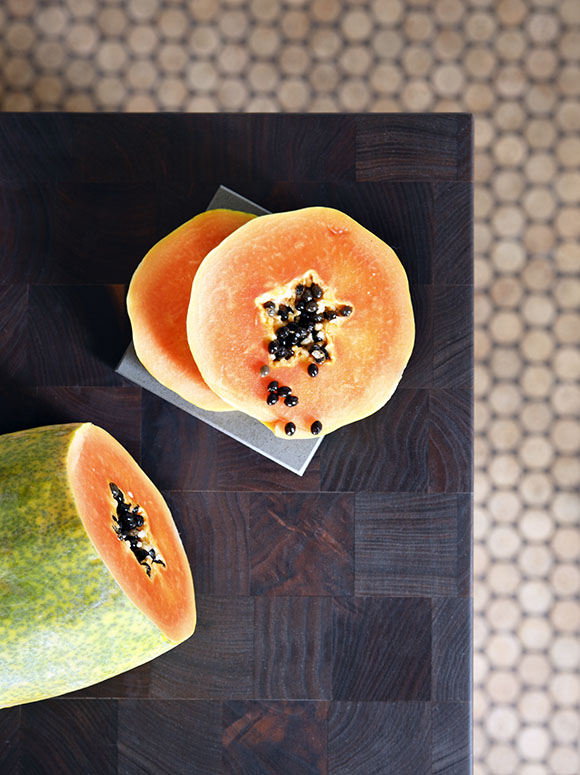 An orange and yellow melon inside with green outsides is sliced open on a butcher block, showing the black seeds inside. In the background, just out of focus, are penny tiles on the floor.