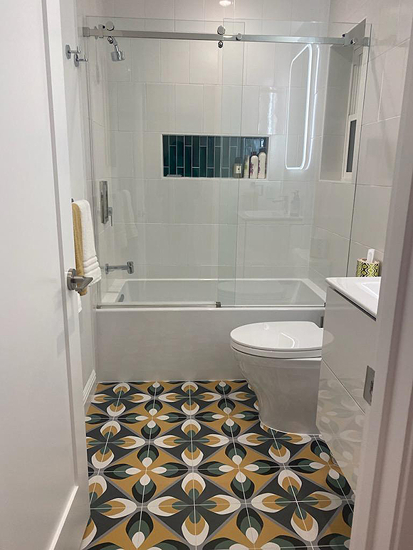 A modern bathroom in mostly all-white: Walls, cabinetry, counters, toilet, and tub with a clear glass shower screen and door. The floor is the most colorful element, being a geographic pattern of squares tilted on their sides in dark blue and grey fields with 4 leaf or feather patterns in white, yellow, and green, connected in the center, alternating with a second pattern of black, green, and white feather shapes. Very hip and mod!