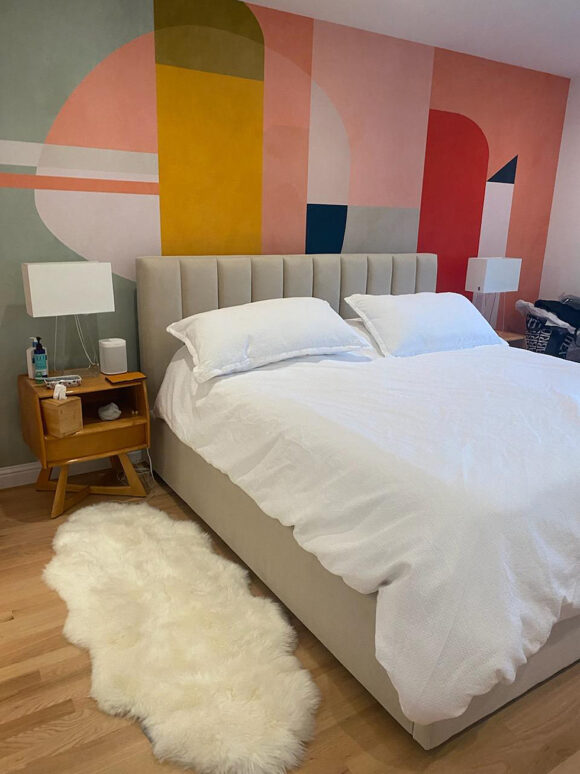A modern bedroom with a graphic mural covering the entire wall at the head of the bed in pink, yellow, blue, red, white, and black geometric shapes overlapping. A queen size bed with a velvet upholstered headboard and matching bed, under white sheets and pillowcases. A small, tan wooden bedside table with a drawer and cubby stands on midcentury modern sideways V-shaped legs. A pair of bedside lamps in clear square glass with square white lampshades sit either side of the bed. On the floor, a faux white animal pelt as a rug for luxurious comfort.