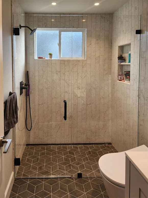 A curbless walk-in shower with a clear glass door showing the walls have an beige and cream textured finish vertical tile, while the floors have a four-sided coppery-brown tile in a geometric shape. The door handle and shower fixtures are black.