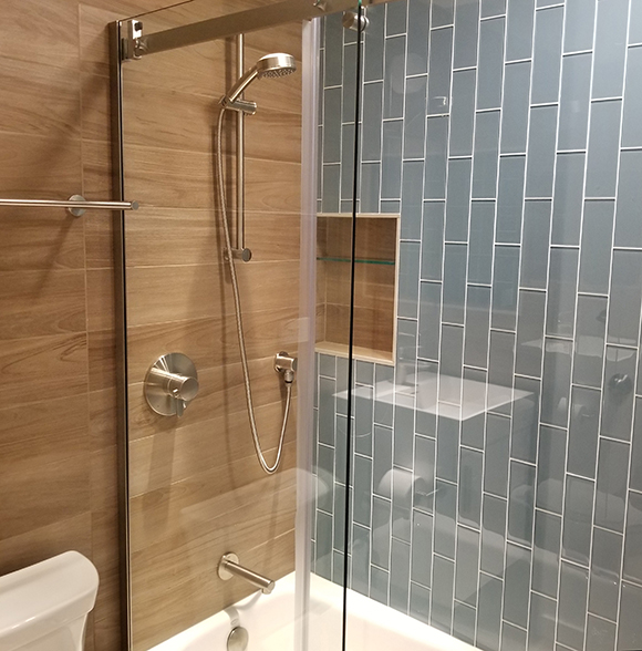 The left side of a bathroom toilet and tub/shower area is tiled with porcelain, wood-look planks on the wall and in the shower wall inset. The right wall is finished in blue-grey vertical glossy tiles.