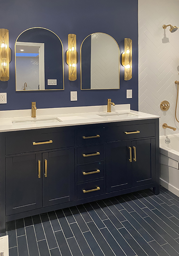 Long blue porcelain floor planks run towards a bathroom counter with two sinks. Three gold wall sconces on a wall painted royal blue with a pair of arched mirrors hang above the gold single faucets at double sinks under white countertops. Dark blue cabinetry with gold handles match the gold shower and tub fixtures over white herringbone tiling and a white tub.