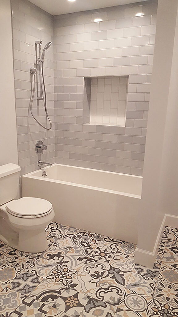 A bathroom composed of off-white, grey, and beige tiles in the shower with a white tub, white toilet, and white tiled shower wall inset. The flooring is a mix of mismatched Moroccan patterned tiles in cornflower blue, tan, mustard, and dark grey.