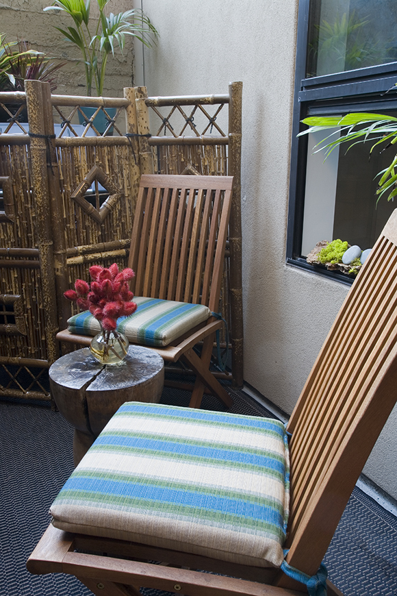 A pair of teak outdoor chairs with blue, green, and white striped seat pads to welcome you. A small round wooden table sits between the two chairs, with a vase of red flowers.