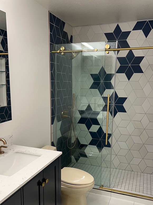 A step-in shower without a tub features large dark blue tiles creating a 6-pointed star surrounded by tan, beige, and off-white tiles in the same shape. The floor is small square off-white tiles. A gold handle on the clear glass door matches the gold sink faucet. The mirror and medicine cabinet are surrounded by the same dark blue tiles as the shower.