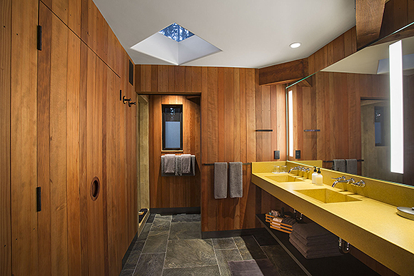 A bathroom clad with redwood. A long yellow countertop holds 2 sinks with soap. Grey towels are layered on two separate racks. Grey ceramic floor tiles stand out underneath a square skylight in the white ceiling. Doors to a sauna and shower can be seen on the left. A huge mirror above the sinks reflects light around the space.