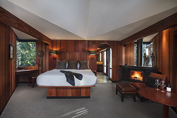 A bedroom view with white angled ceilings above redwood-clad walls, an arched entrance to the bathroom, angled windows, one with a desk in front, a fireplace, a wooden table dressed with wine glasses and a bottle, a leather footstool by the fire, and a king size platform bed on matched redwood, with wall sconce bedside lamps at the head. The floor is grey.