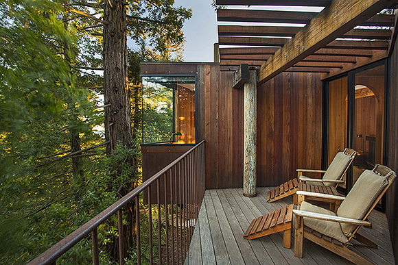 Outside on a wooden balcony of the treehouse, with 2 adirondack-style loungers with seat and back pads in grey are facing out into the forest. A glass corner window is visible to the side, while exposed beams are held up by a wooden pole that rises through the wood floor. A metal rail extends towards you.