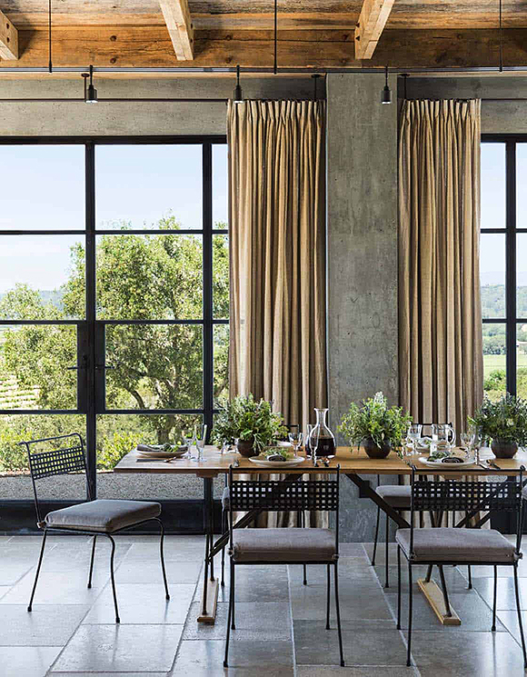 A very high ceiling of exposed wood beams warms up a wine tasting space created by metal window frames, looking out to a view of green trees and orchards. Natural off-white curtains are gathered next to a concrete post on the sides of the windows. Oversized floor tiles in bronze with darker patina support a dining set on delicate black metal legs, which a wooden top on the rectangular dining table, dressed with 3 vases of greens, a flask full of wine, and table settings for 5 that we can see -- the end of the table on the right is cut off by the camera. One chair is pulled out as if ready to welcome you.