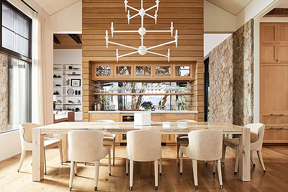An open dining room with a built-in custom bar for wine tasting, and a high ceiling. The room is designed in off-whites, beiges, copper colors, and warm cherry wood. 8 off-white leather dining chairs from Roche Bobois surround a reclaimed wood rectangular dining table. A white, tiered, mod-style plaster light fixture from Bourgeouis Boheme counters the height of the ceilings and brings sophistication and lightness to the dining space.