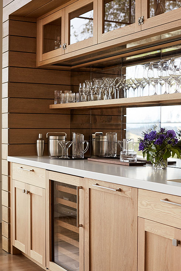 A closeup view of the bar, with white oak wood custom cabinetry below and patina-mirrored tiles above reflecting the vineyard, with white oak open shelves and glass door cabinets above the counter. A wine chiller is visible through the glass door underneath the counter, between cabinet doors, one concealing a mini dishwasher.