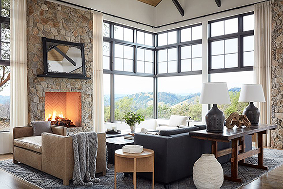 A tall, corner aspect window with a view out to the mountains and vineyards frames a comfortable living room with a stone fireplace and stone walls extending up towards exposed beams and a white ceiling. A dark blue sofa sits at a 90-degree angle to a double-sided leather bench seat. Behind the blue sofa is a console table with a pair of blue table lamps and white lampshades. An antique mirror sits above the fireplace without a mantel, on a shelf.